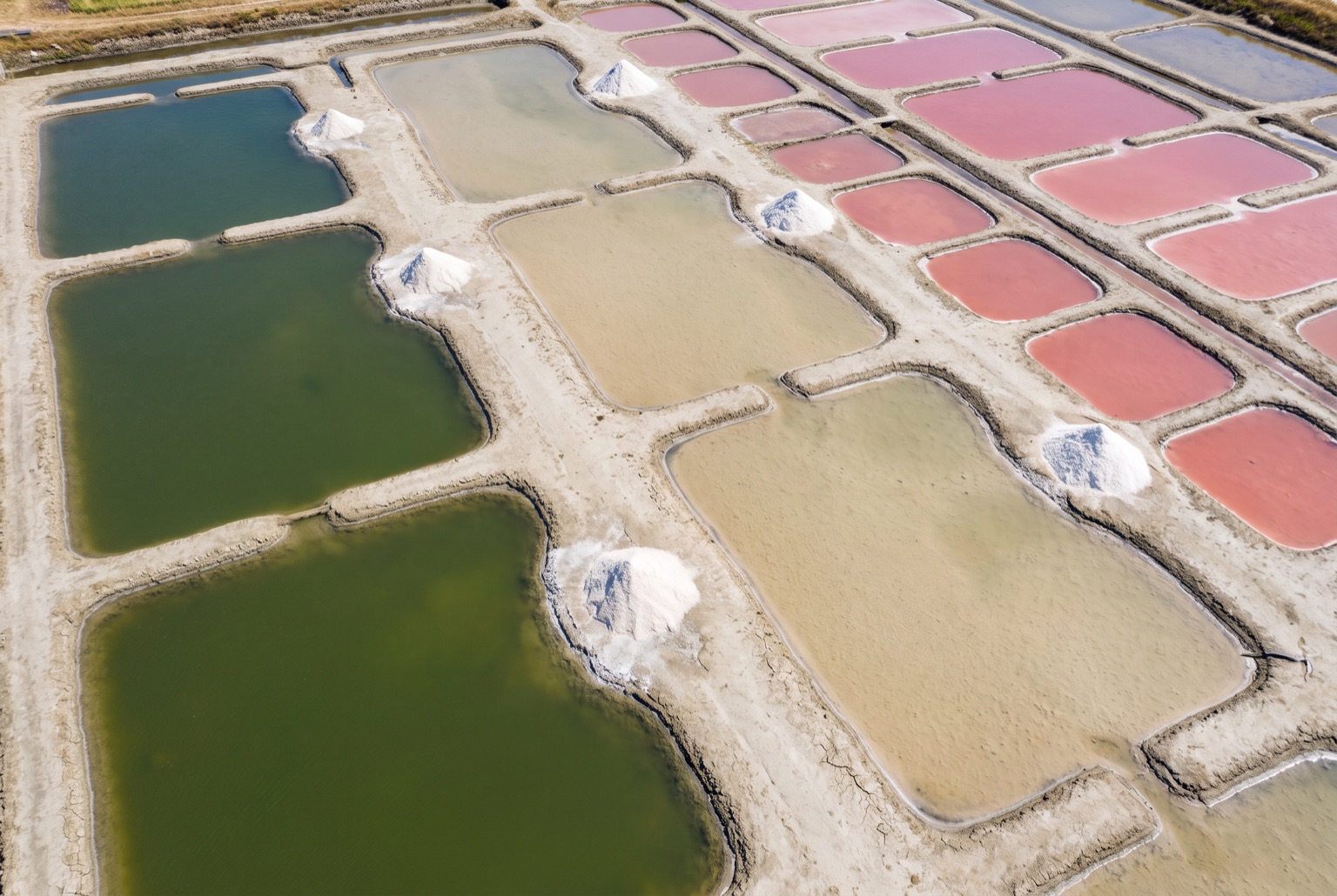 The saline from above: shallow clay pans in a hand-cut grid, the water deep green in the largest, pale sand in the middle ones and vivid rose in the smallest, with white heaps of harvested salt on the banks.